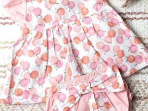 Two piece baby girls romper set. A peach design printed top with light pink ruffled caped shoulders and a diaper cover with a large bow on a neutral background.