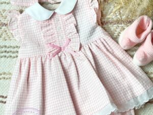 Round collar baby dress with checkered design and ruffles. Three little buttons on the chest and a tiny pink bow.  Paired with knicker and bonnet on a neutral background.