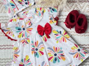 Baby girl white dress with splashes of colors of red teal navy blue yellow orange and pink with a ruffled collar and capped sleeve with a bow on a neutral background.