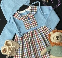 A light blue combination knit checkered dress set with a blue bonnet. Has a peter pan collar with checkered print decorated with two buttons on a black backdrop. Tan newborn knit baby booties and stuffed lion toy in the photo.