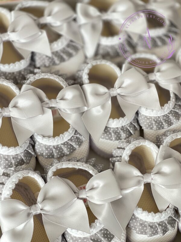 Several grey and white satin baby shoes with a polkadot ruffle trim and bow lined up in rows.