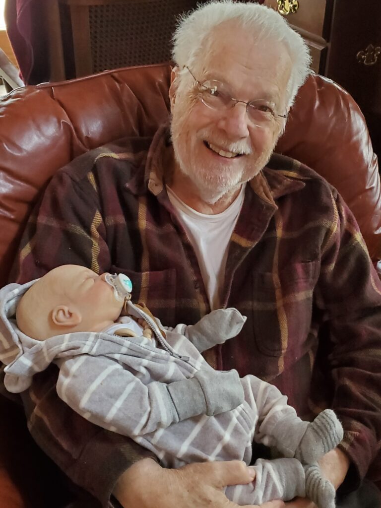 A special grandpa wearing a red plaid button down shirt with a white t-shirt sitting in a brown leather chair holding a reborn therapy doll